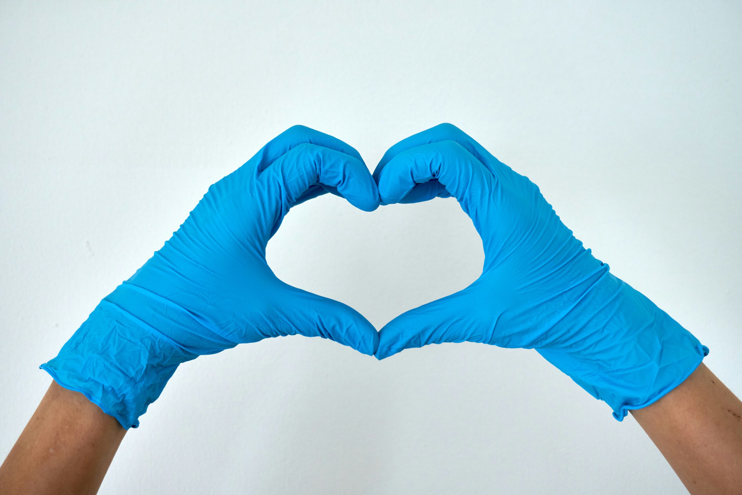 Zu sehen sind ein Paar Hände mit blauen Latexhandschuhen vor einem weißen Hintergrund. Die Hände bilden mit den Fingern ein Herz, wobei die Daumen die untere Spitze bilden. Die gebräunten Unterarme des Models sind ebenfalls zu erkennen.