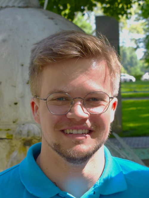 Der Mitgründer Akim Rennette. Ein junger Mann Mitte 20 mit blonden, kurzen Haaren, kurzem Bart und einer silbernen, schmalen Brille. Er trägt ein hellblaues Poloshirt des Unternehmens und lächelt breit. Im Hintergrund sind grüne Bäume, ein Stück blauer Himmel und eine Hausecke, die verschwommen sind, damit die Person besser raussticht.