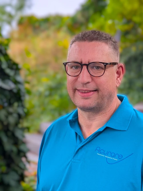 Abgebildet ist der Mitarbeitende Frank Lexa. Ein Mann Mitte 50 mit kurzem grauem Haar, einer schwarzen Brille und einem freundlichen Lächeln. Er trägt ein blaues Poloshirt mit dem Logo und der Aufschrift des Unternehmens: 'Acurare Häuslicher-Assistenz-Dienst'. Im Hintergrund sind grüne Büsche und Bäume und ein grau-brauner Weg abgebildet, die verschwommen sind, damit die Person hervorsticht.