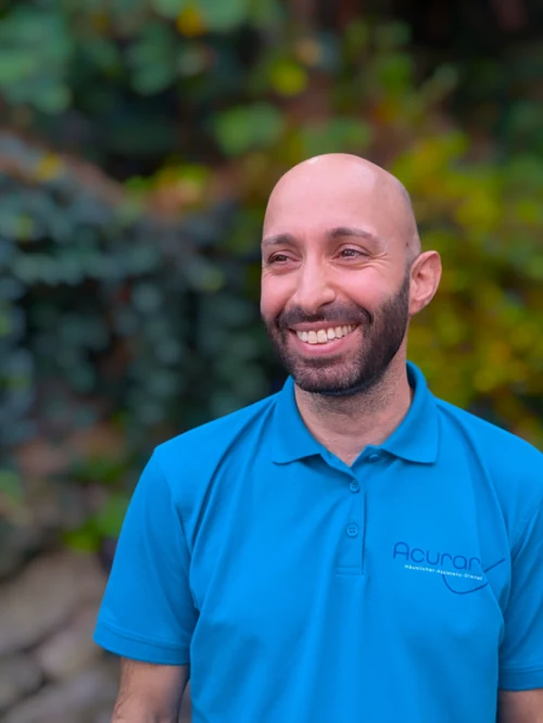 Zu sehen ist der Mitarbeitende Giuseppe Russo. Ein Mann Anfang 40 mit Glatze, gepflegtem, kurzem dunklen Bart und einem breiten Lächeln. Er trägt ein blaues Poloshirt mit dem Logo und der Aufschrift des Unternehmens: 'Acurare Häuslicher-Assistenz-Dienst'. Im Hintergrund sind grüne Büsche und Bäume abgebildet, die verschwommen sind, damit die Person hervorsticht.