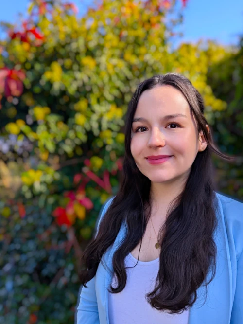 Abgebildet ist die Mitarbeitende Miriam Solano. Sie ist rechtsbündig auf dem Bild, mit langen, dunklen Haaren, lächelnd. Sie trägt ein weißes Oberteil und darüber einen hellblauen Blazer. Um den Hals ist eine Goldkette mit einem kleinen runden Anhänger. Der Hintergrund sind verschwomene grüne und rote Büsche.