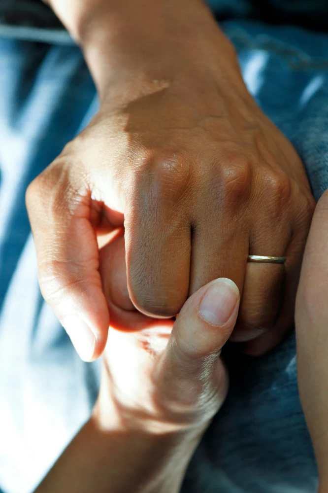 Abgebildet sind zwei Paar Hände und ein Bein in Jeansstoff im Hintergrund. Eine Hand von links umfasst liebevoll die rechte Hand mit Ehering einer anderen Person in Jeans. Am unteren Bildrand ist die andere Hand der Person von links zu erkennen. Die Sonne scheint auf die Hände.