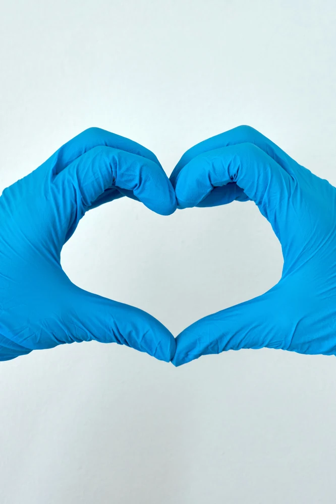 Auf dem Bild ist ein Paar Hände abgebildet auf einer weißen Fläche. Die Hände stecken in blauen Latexhandschuhen. Die linke Hand wischt mit einem Küchenpapier die Fläche, die rechte Hand umfasst eine Sprühflasche. Die Sprühflasche hat einen dunkelblauen Sprühkopf, ein grün-weißes Etikett der Marke Dettol.